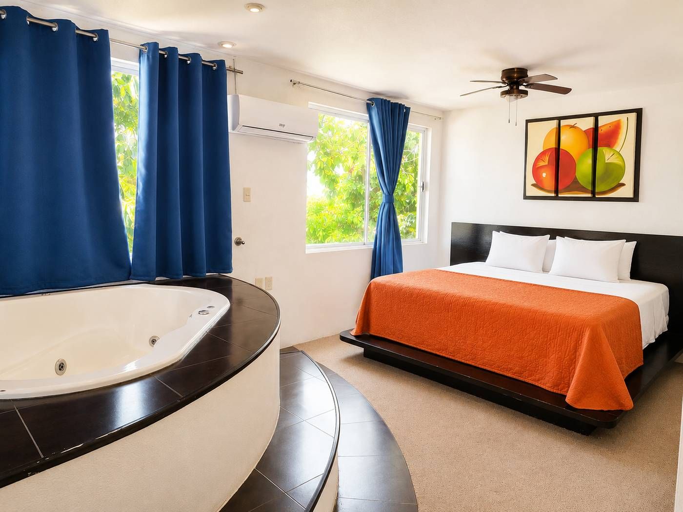 Quarto de hotel com banheira curva, cama com colcha laranja, ventilador de teto e janela com cortinas.