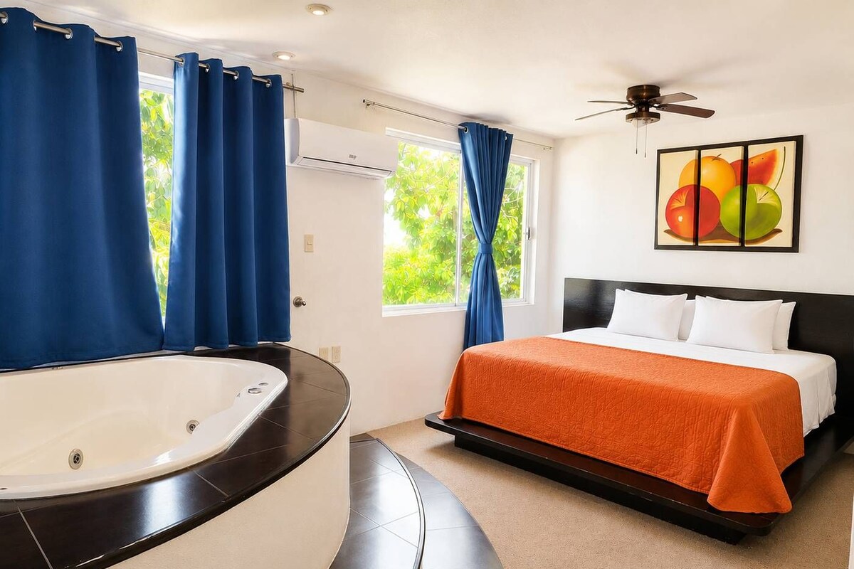 A hotel room with a curved bathtub, a bed with an orange bedspread, a ceiling fan, and a window with curtains.