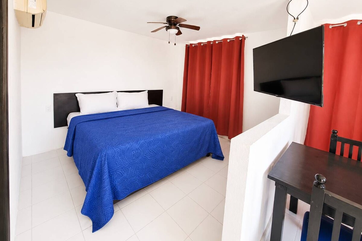 A hotel room with a bed, a desk, a TV, a ceiling fan, and red curtains.