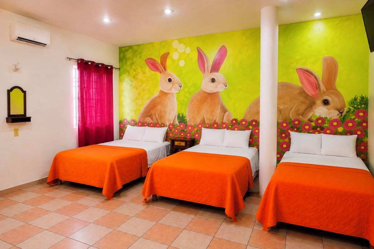 A hotel room with three beds, each with an orange bedspread, a large wall mural of rabbits, and a red curtain.