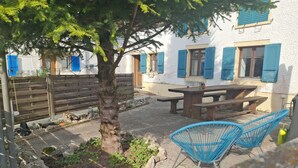 En terrasse med et træbord og stole, et træ og en hvid bygning med blå skodder.