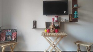 A room with a flat-screen TV mounted on the wall, a small wooden table with a few items on it, two folding chairs, and a shelf with decorative objects. - Charming Duplex in enchanting Marrakesh with cool AC (Marrakesh)