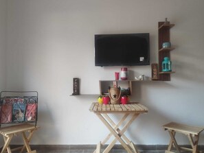 Ein Zimmer mit einem an der Wand befestigten Flachbildfernseher, einem kleinen Holztisch mit einigen Gegenständen darauf, zwei Klappstühlen und einem Regal mit Dekoration.
