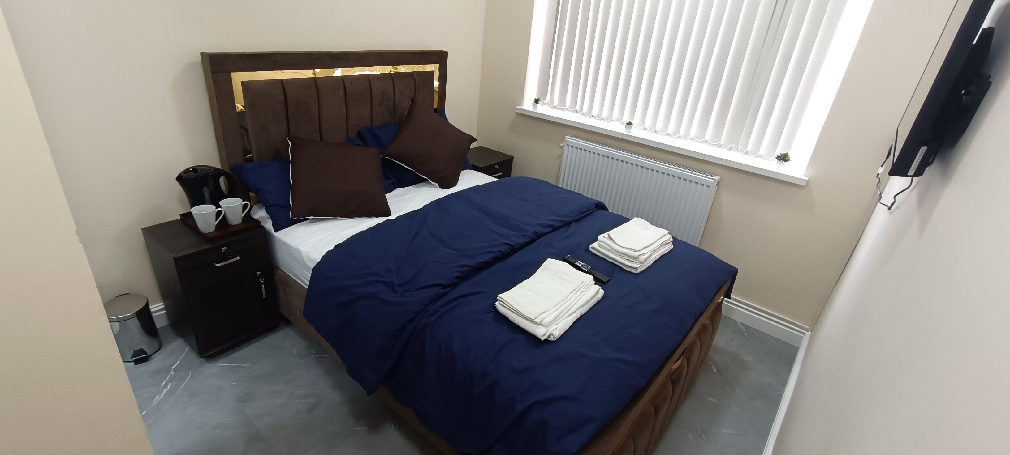 A neatly made bed with a blue bedspread and pillows, a bedside table with a kettle and cups, and a window with blinds.