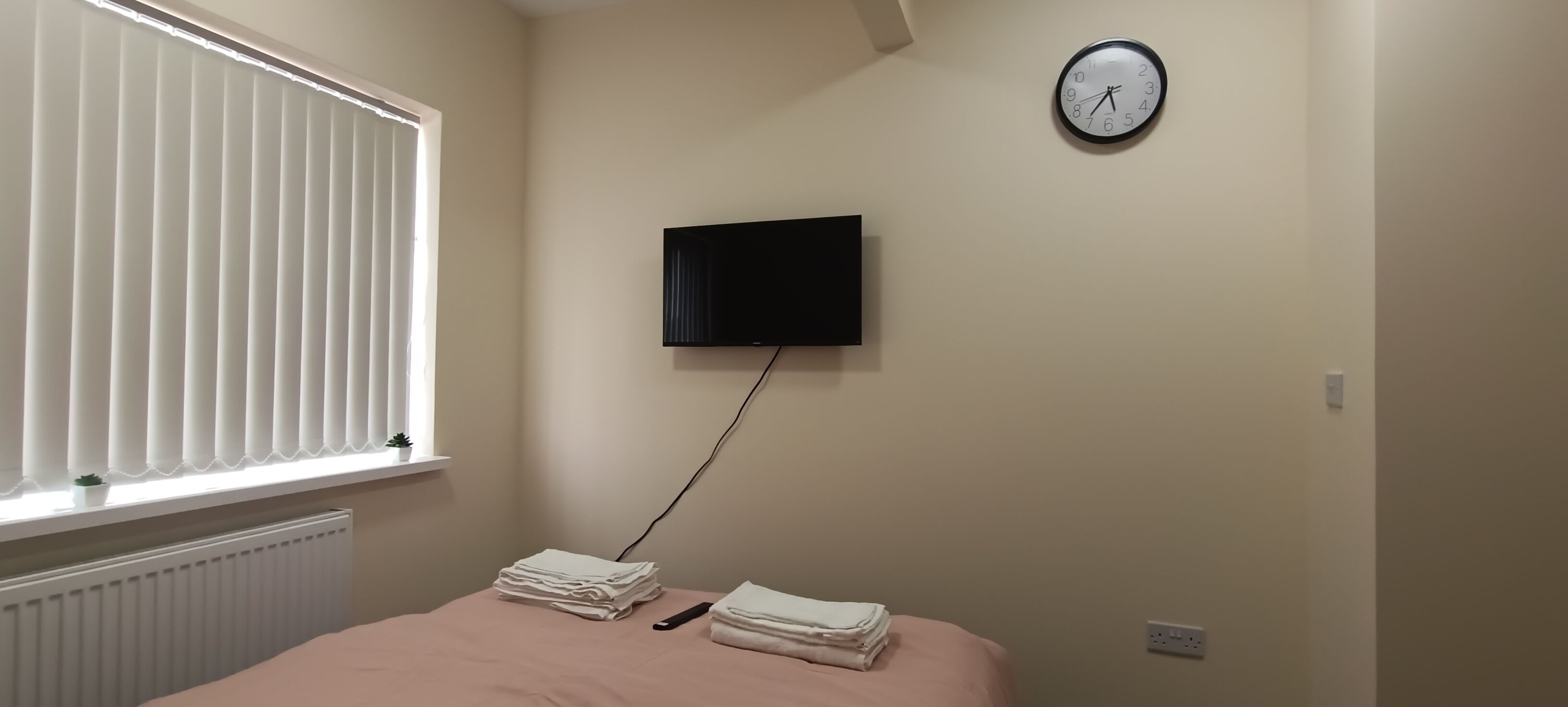 A bedroom with a bed, a television mounted on the wall, a window with blinds, and a radiator.