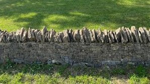 Ein dreieckiges Haus mit sichtbarem Balkon, umgeben von einer Steinmauer und Grünpflanzen.