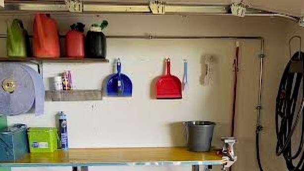 Eine Garage mit Werkbank, Regalen voller Werkzeuge und Behälter, ein grüner Rasenmäher und eine Gartenschlauchaufroller.