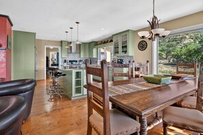Un coin repas avec une table et des chaises en bois, une cuisine à l’arrière-plan et une vue sur l’extérieur.