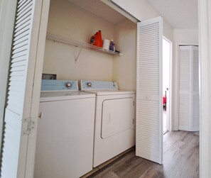 A laundry room with two washing machines, a shelf with cleaning supplies, and a door leading to another room. - Close Disney! Spacious 3Br 2 Bh Condo / 75" TV, Pool, Parking & No I-4 Traffic! (Kissimmee)