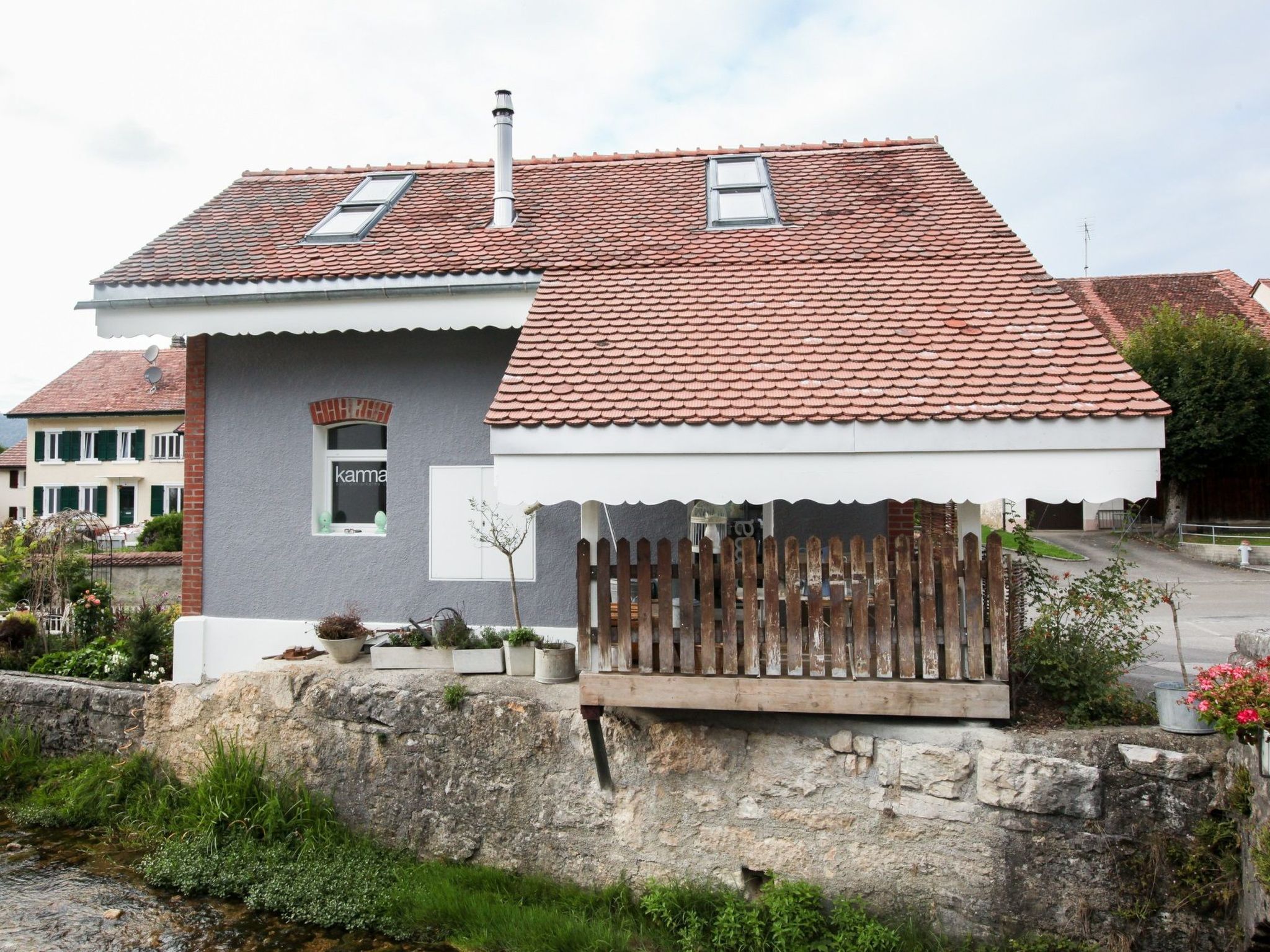 Una casa de dos pisos con techo de tejas rojas, un pequeño jardín y un muro de piedra.
