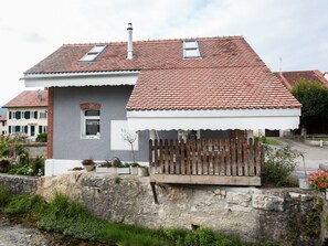 Una casa de dos pisos con techo de tejas rojas, un pequeño jardín y un muro de piedra.