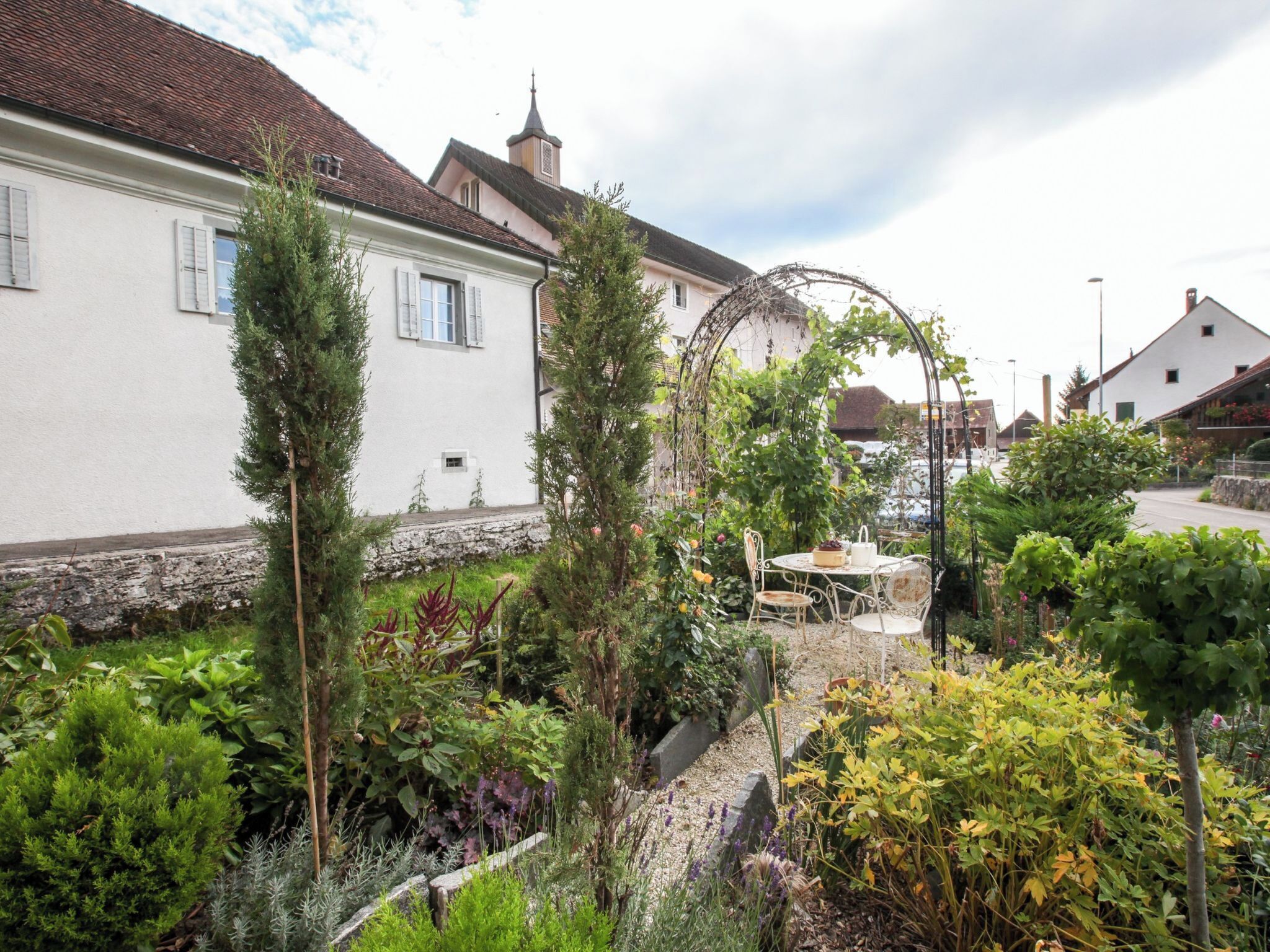A garden with a table and chairs, surrounded by various plants and a white building with a red roof.