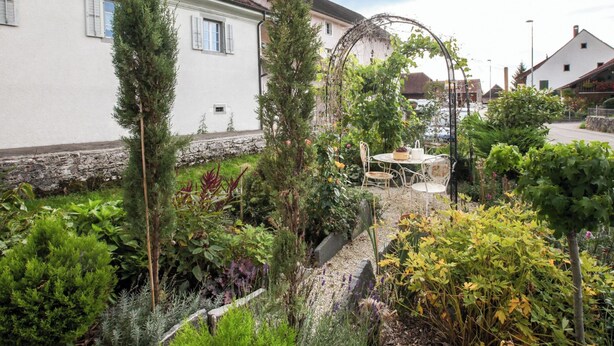 Ein Garten mit Tisch und Stühlen, umgeben von Pflanzen und einem weißen Gebäude mit rotem Dach.