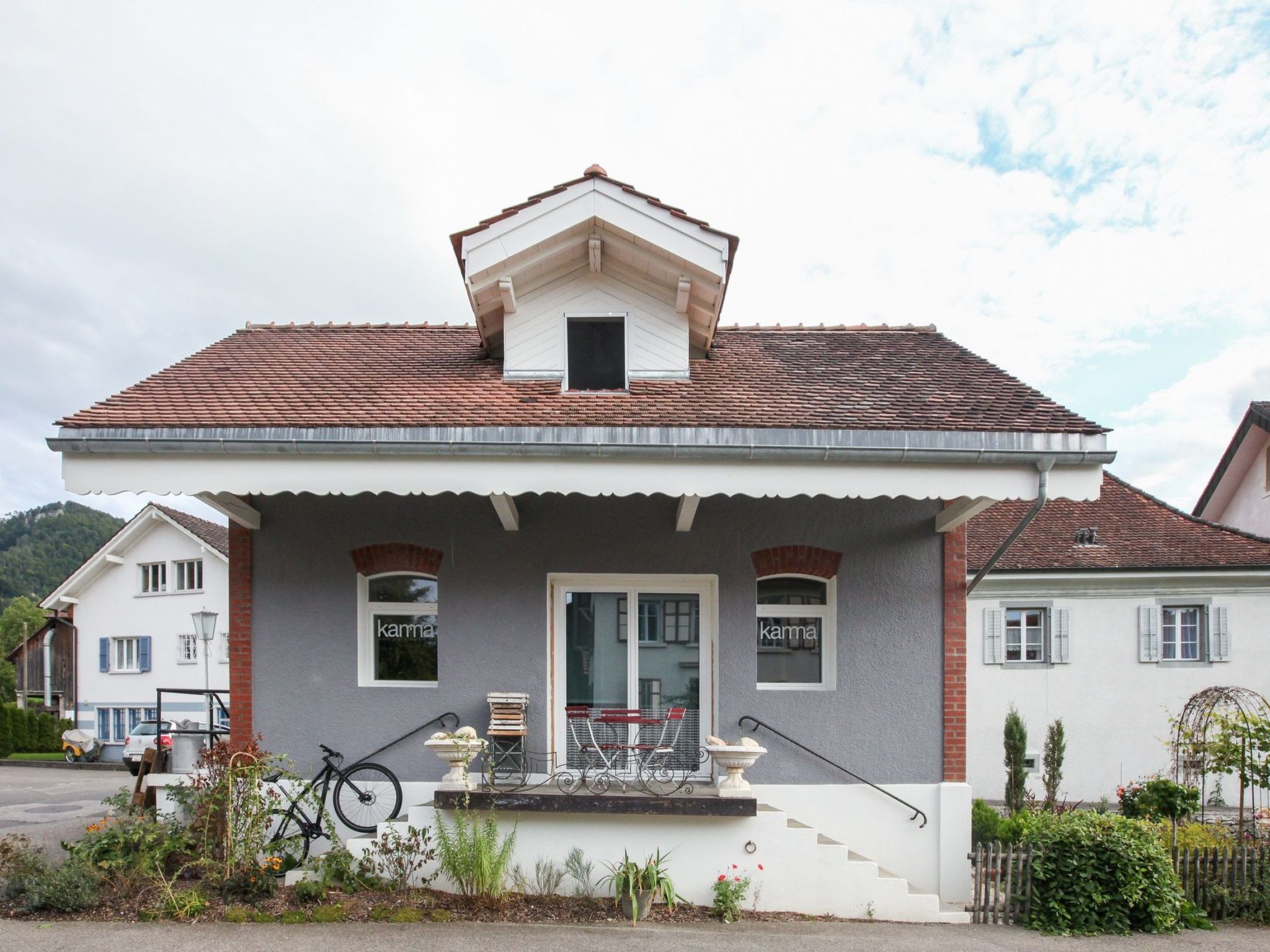 A two-story house with a tiled roof, a front porch with a bicycle, and a small window with the word 'kama' on it.