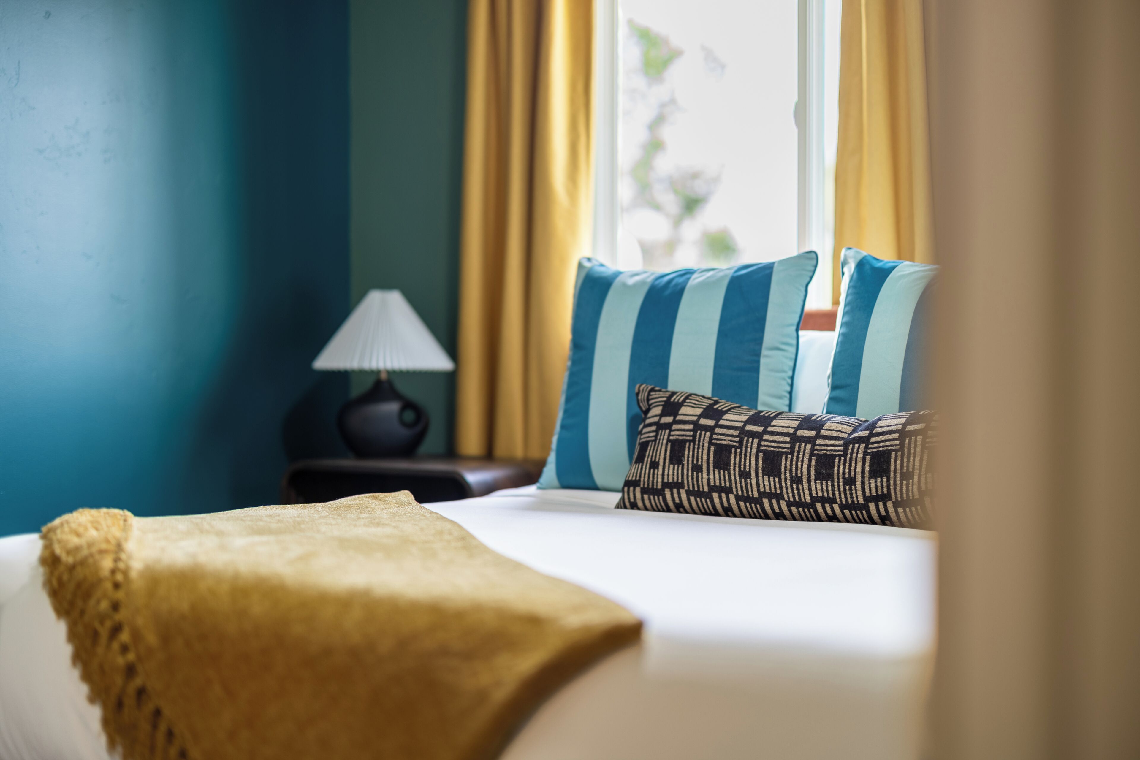 Una cama bien tendida con un edredón amarillo, dos almohadas con diseños a rayas y estampados, y una lámpara en una mesita de noche.