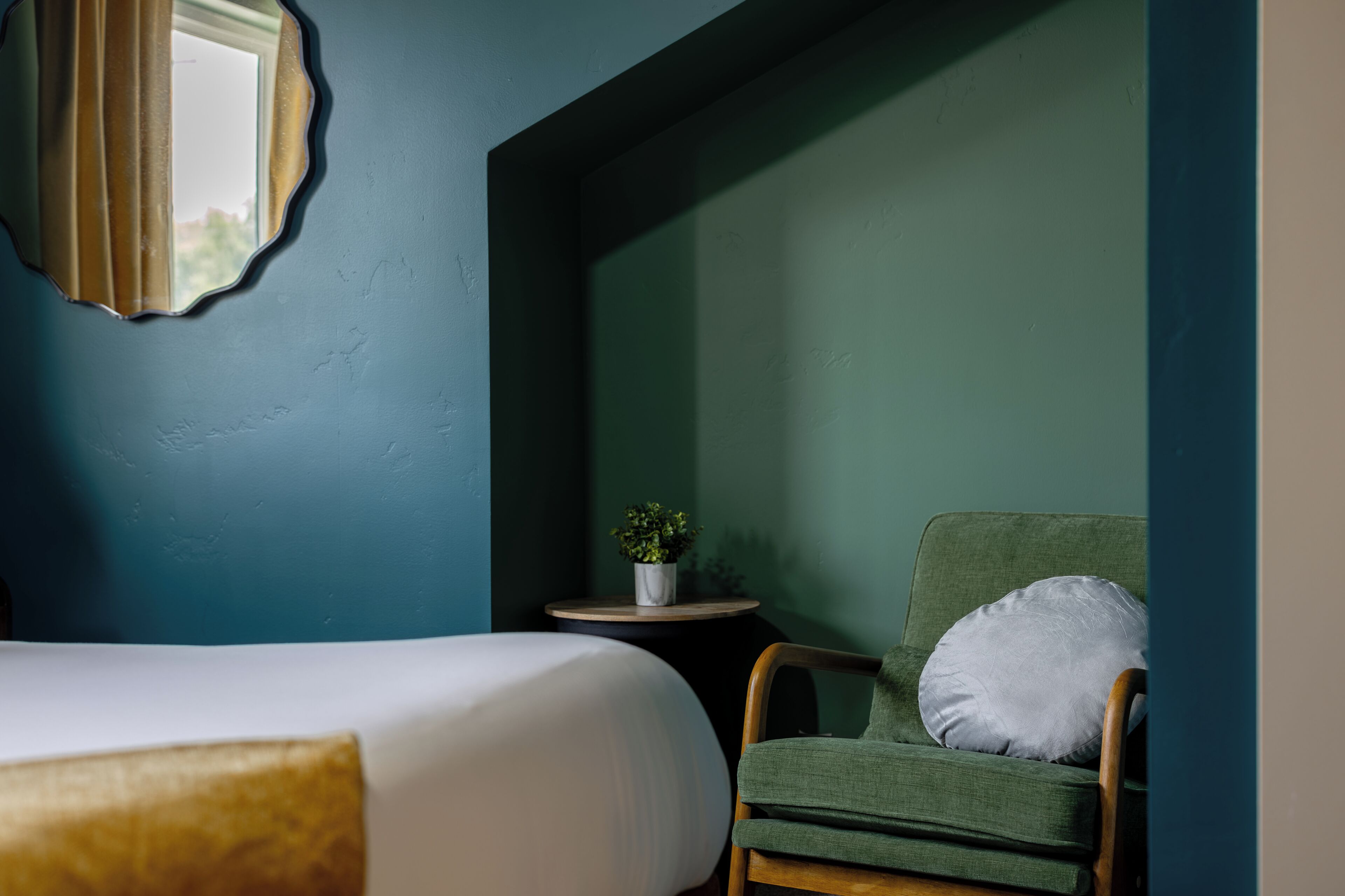 A room with a bed, a chair with a pillow, a small table with a plant, and a mirror on the wall.