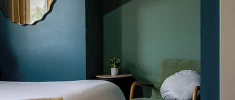 A room with a bed, a chair with a pillow, a small table with a plant, and a mirror on the wall.