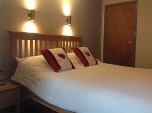 Una cama bien hecha con ropa blanca y dos almohadas rojas, un cabecero de madera y dos lámparas fijadas a la pared.