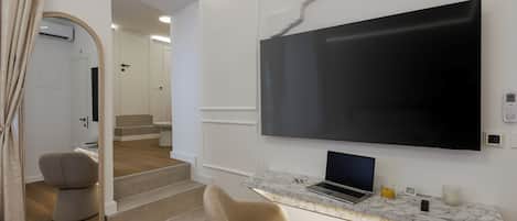 Un salon moderne avec un téléviseur à écran plat fixé au mur, un comptoir en marbre sur lequel se trouve un ordinateur portable, un fauteuil gris et un escalier menant à une autre pièce.