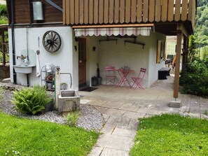 Een overdekt terras met een stenen vloer, een rustieke houten pergola en een kleine tuin met varens en grind.