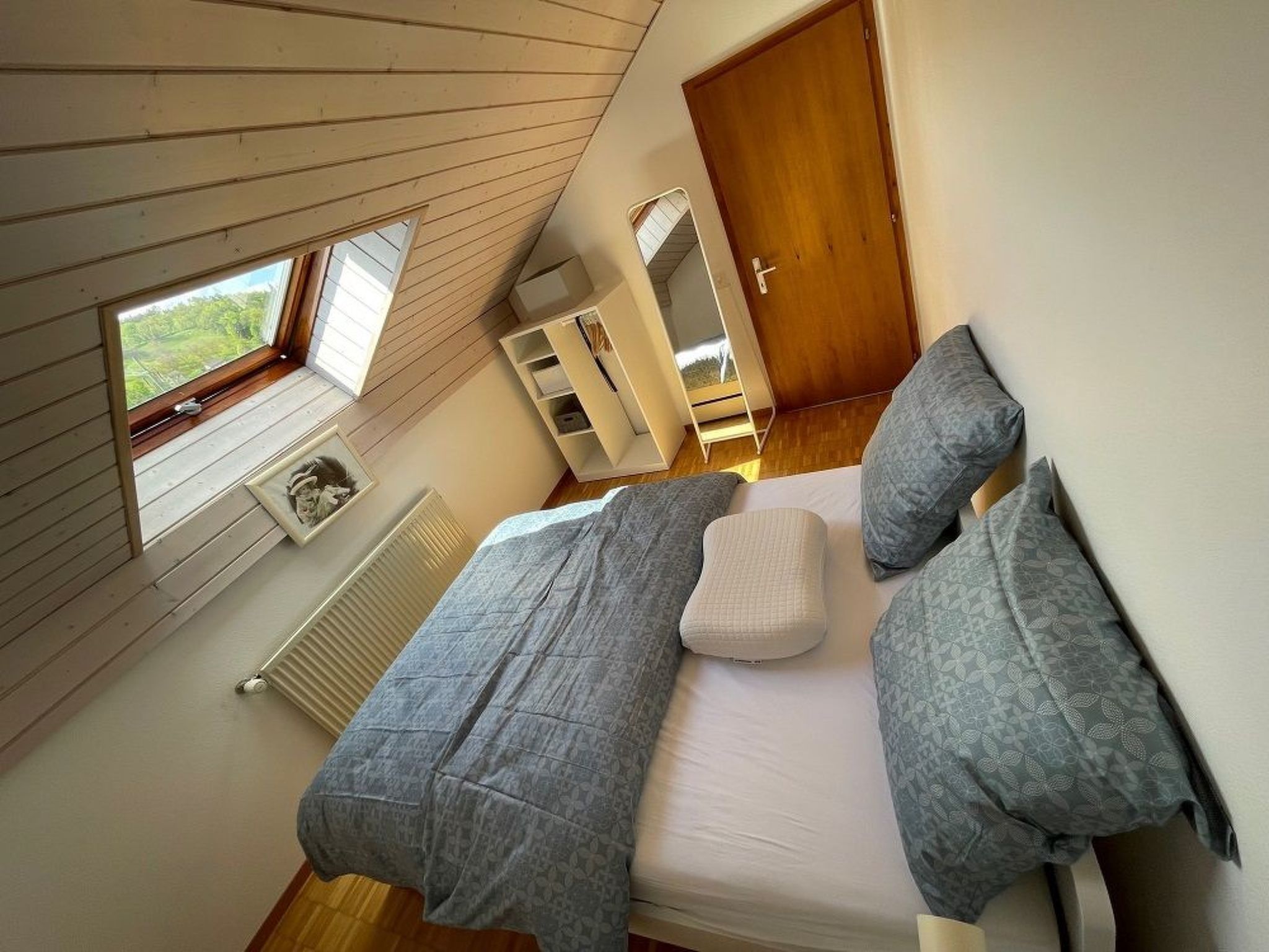 Un dormitorio con techo inclinado, cama con ropa de cama gris, ventana y radiador.