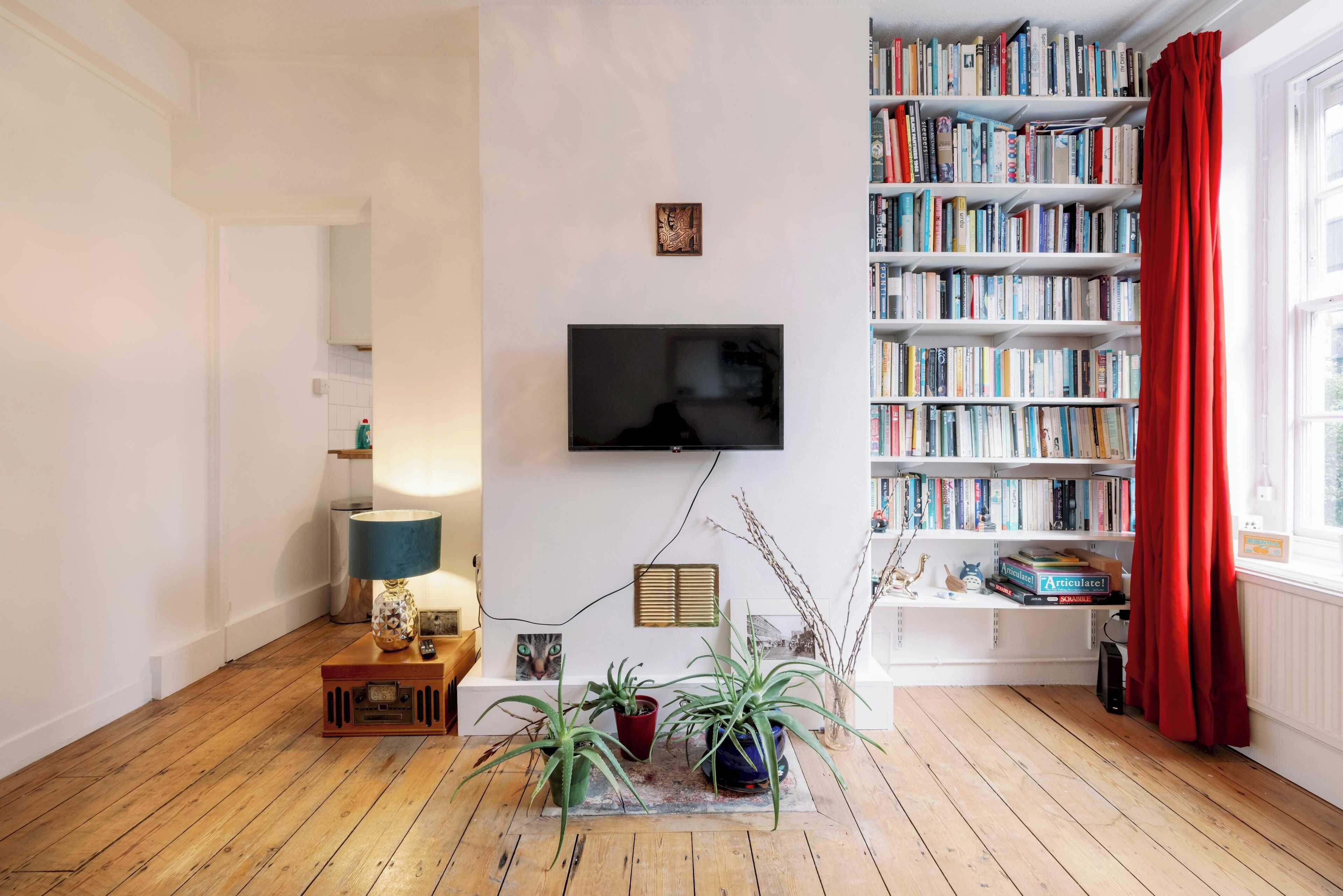 Ein Wohnzimmer mit Holzboden, einem an der Wand befestigten Flachbildfernseher, einem Bücherregal voller Bücher und einem roten Vorhang am Fenster.
