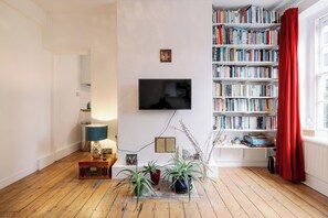 A living room with a wooden floor, a flat-screen TV mounted on the wall, a bookshelf filled with books, and a red curtain on the window. - Stylish 1-Bed in Bethnal Green • Calm & Central - Pass the Keys (London)