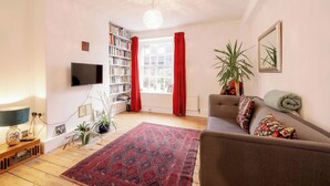 A living room with a television, bookshelf, red curtains, a sofa, and a patterned rug. - Stylish 1-Bed in Bethnal Green • Calm & Central - Pass the Keys (London)