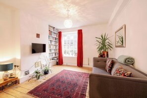 A living room with a television, bookshelf, red curtains, a sofa, and a patterned rug. - Stylish 1-Bed in Bethnal Green • Calm & Central - Pass the Keys (London)