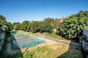 A fenced sports court with a basketball hoop, surrounded by trees and a brick building in the background. - Stylish Leith Flat | Great Transport Links to City - Pass the Keys (Edinburgh)