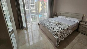 Habitación de hotel con cama, mesita de noche, ventana con cortinas y vistas a edificios.