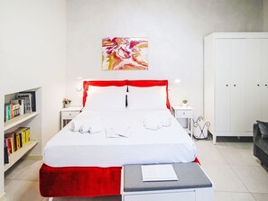 A neatly made bed with white linens and red accents, a bedside table with a book and a small plant, a bookshelf with various books, and a large abstract painting on the wall. - Beautiful apartment for 3 people with WIFI, A/C, TV and balcony (Colazza)