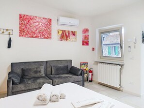Ein modernes Zimmer mit einem grauen Sofa, einem Bett mit aufgerollten Handtüchern und einem Fenster mit Blick auf ein Gebäude.