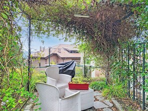 Un espace extérieur aménagé avec des chaises en osier et un jardinier rouge, entouré de verdure et offrant une vue sur une maison.