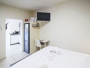 Ein kleines, sauberes Zimmer mit einem Bett, einem Schreibtisch, einem Stuhl, einem Kühlschrank und einem an der Wand befestigten Fernseher.