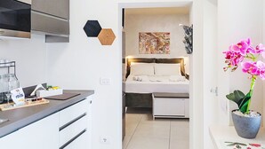 A compact hotel room with a kitchenette, a bed with a headboard, a painting on the wall, and a potted orchid. - Beautiful apartment for 3 guests with WIFI, A/C, TV and balcony (Colazza)
