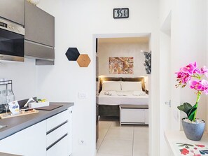 A compact hotel room with a kitchenette, a bed with a headboard, a painting on the wall, and a potted orchid.