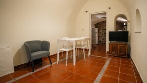 A room with a sofa, a dining table with chairs, a television on a cabinet, and a stone wall in the background.