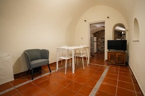 A room with a sofa, a dining table with chairs, a television on a cabinet, and a stone wall in the background.