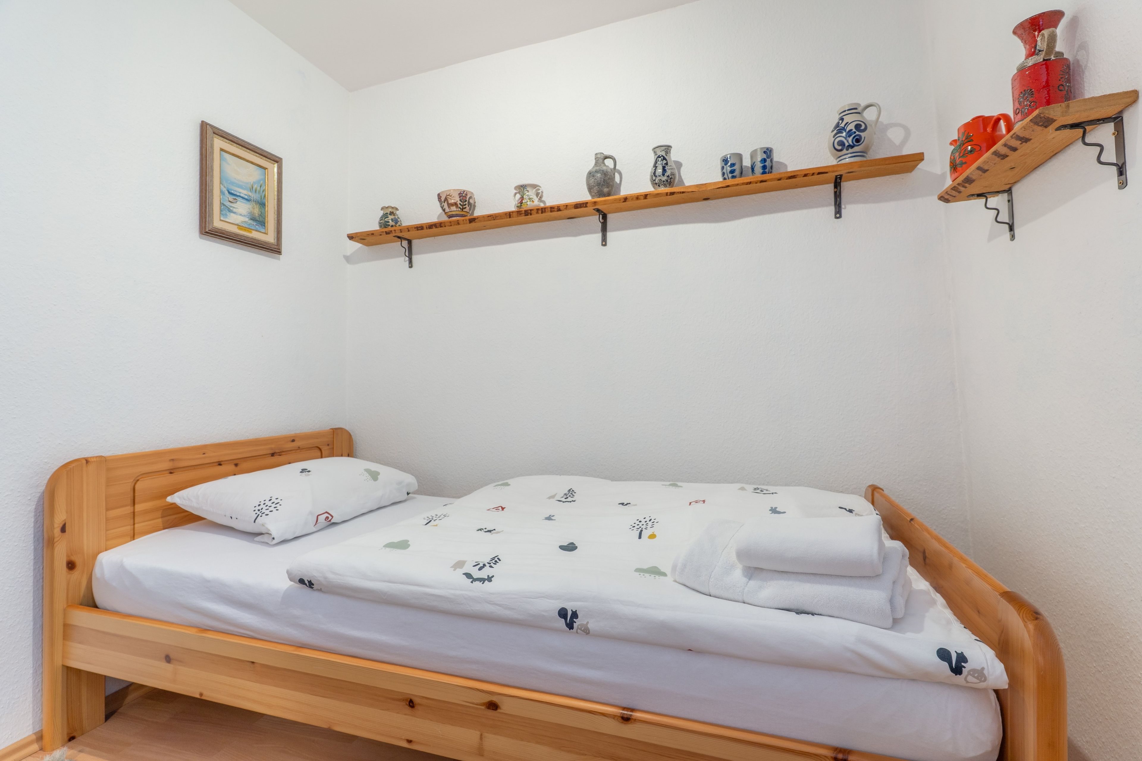 A single bed with a wooden frame and white bedding, a shelf with decorative items, and a framed picture on the wall.