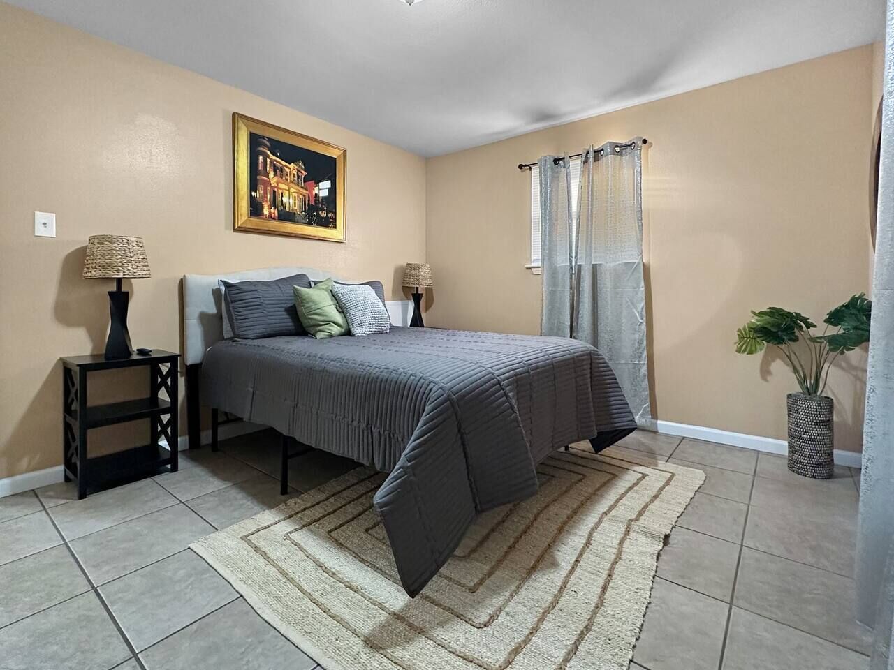 Une chambre à coucher avec un lit, des tables de chevet, un tableau, une fenêtre avec des rideaux et une plante.