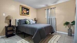 Un dormitorio con cama, mesitas de noche, un cuadro, una ventana con cortinas y una planta.