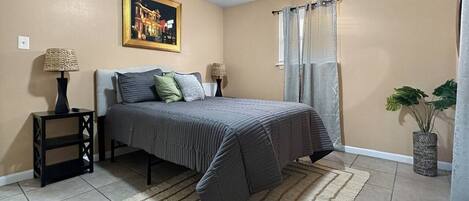 A bedroom with a bed, nightstands, a painting, a window with curtains, and a plant.