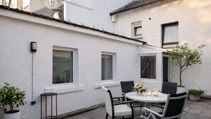 A patio with a round table and chairs, potted plants, and a white wall with windows.