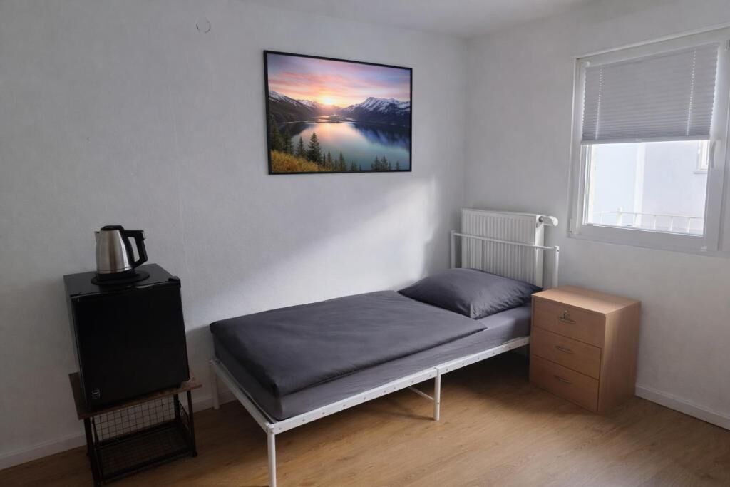 Une petite chambre avec un lit simple, donnant sur un lac et des montagnes, équipée d’un radiateur, d’une fenêtre avec stores, d’une petite armoire et d’un chauffage sur pied.