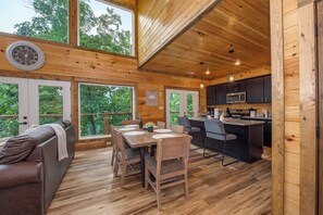 L’intérieur d’un chalet en bois comprend un coin repas, une cuisine et une grande fenêtre donnant sur un paysage verdoyant.