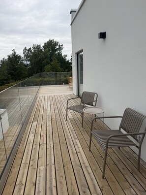 Eine Holzterrasse mit Metallstühlen und einem Tisch, mit Blick auf ein bewaldetes Gebiet.