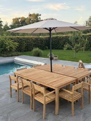 Ein Holztablett mit Stühlen und einem großen Sonnenschirm, vor einer Pool- und Gartenlandschaft.