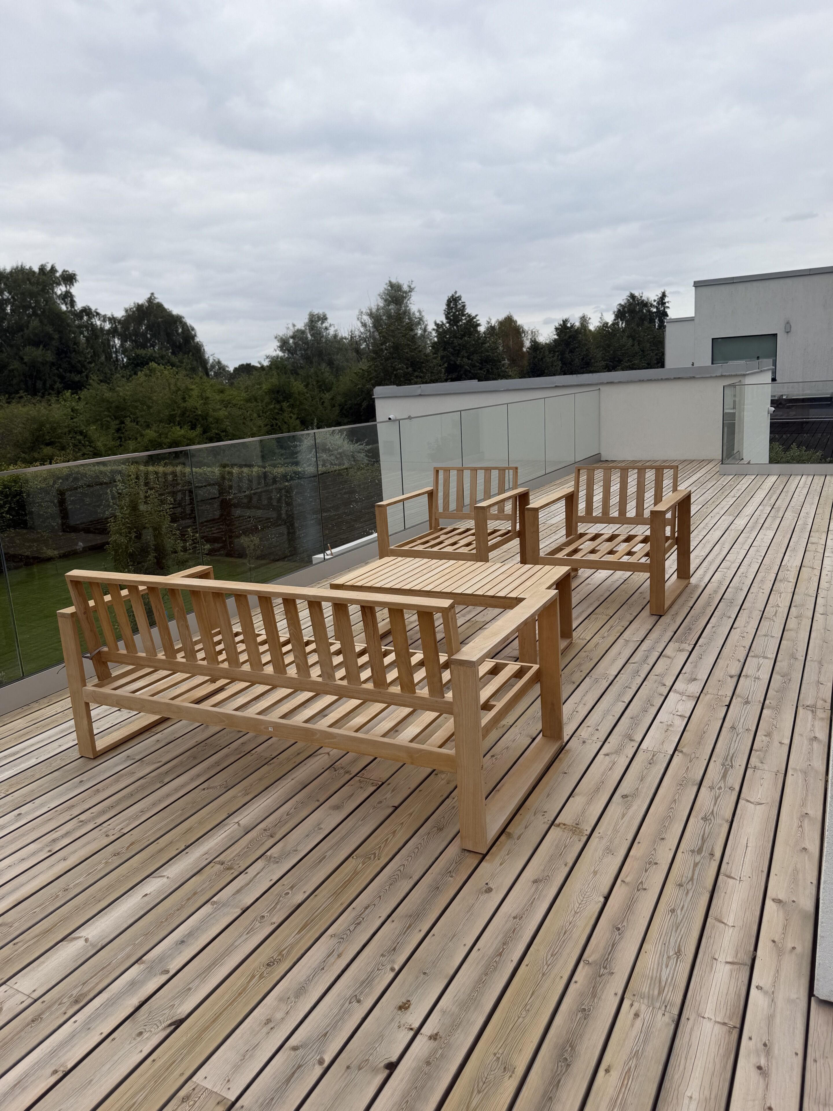A wooden deck with a bench and a table, surrounded by a glass railing, overlooking a green landscape.