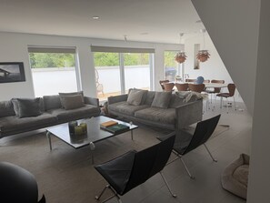 Ein modernes Wohnzimmer mit einer Couch, einem Couchtisch und Stühlen, großen Fenstern und einem Essbereich mit Tisch und Stühlen.